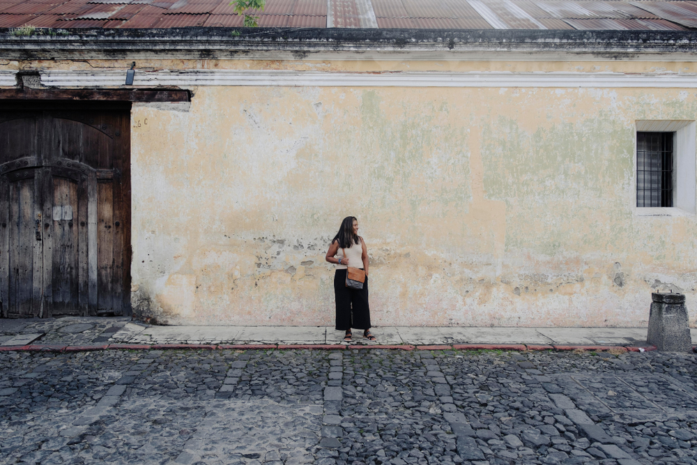 A woman stands on a cobblestone street in front of a large, weathered beige wall with a dark wooden door on the left. She is wearing a light beige top, a black skirt, and carrying a brown shoulder bag.