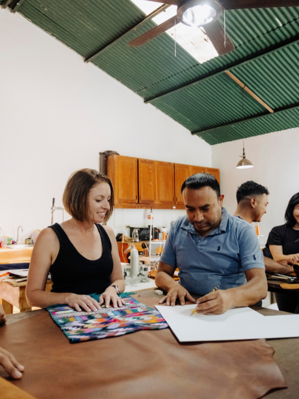 A man and a woman sit at a table in a workshop, working on craft projects. The woman holds a colorful geometric patterned fabric, while the man writes on a white sheet of paper.