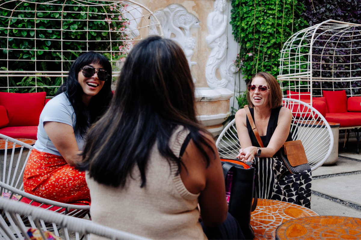 Three women laughing and sitting in a relaxed outdoor lounge setting with white woven chairs, red cushions, and greenery in the background.