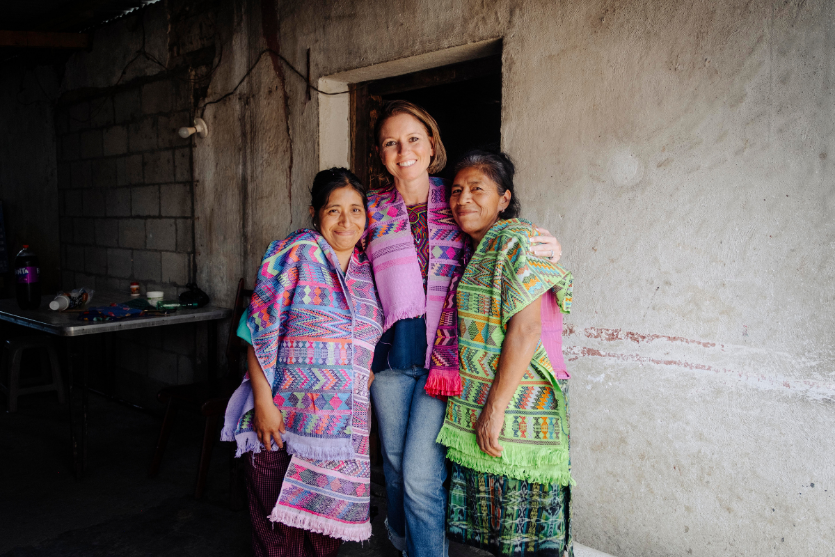 Three women standing together in front of a rustic concrete wall doorway, wearing colorful woven Peruvian-style textiles in pink, green, blue, and purple.