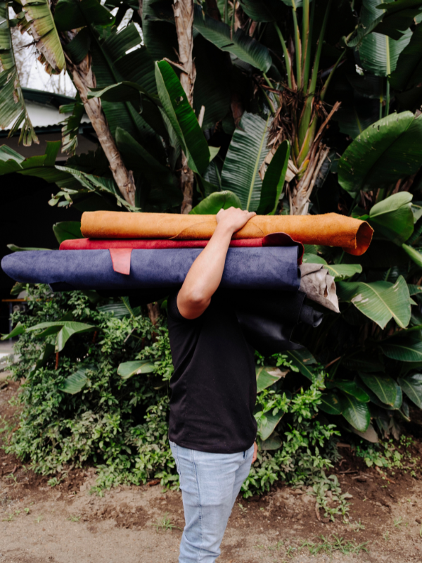 A person outdoors carrying three rolled exercise mats on their shoulder. The mats are stacked horizontally, showing different colors: a tan/orange mat on top, a red mat in the middle, and a dark blue mat on the bottom.