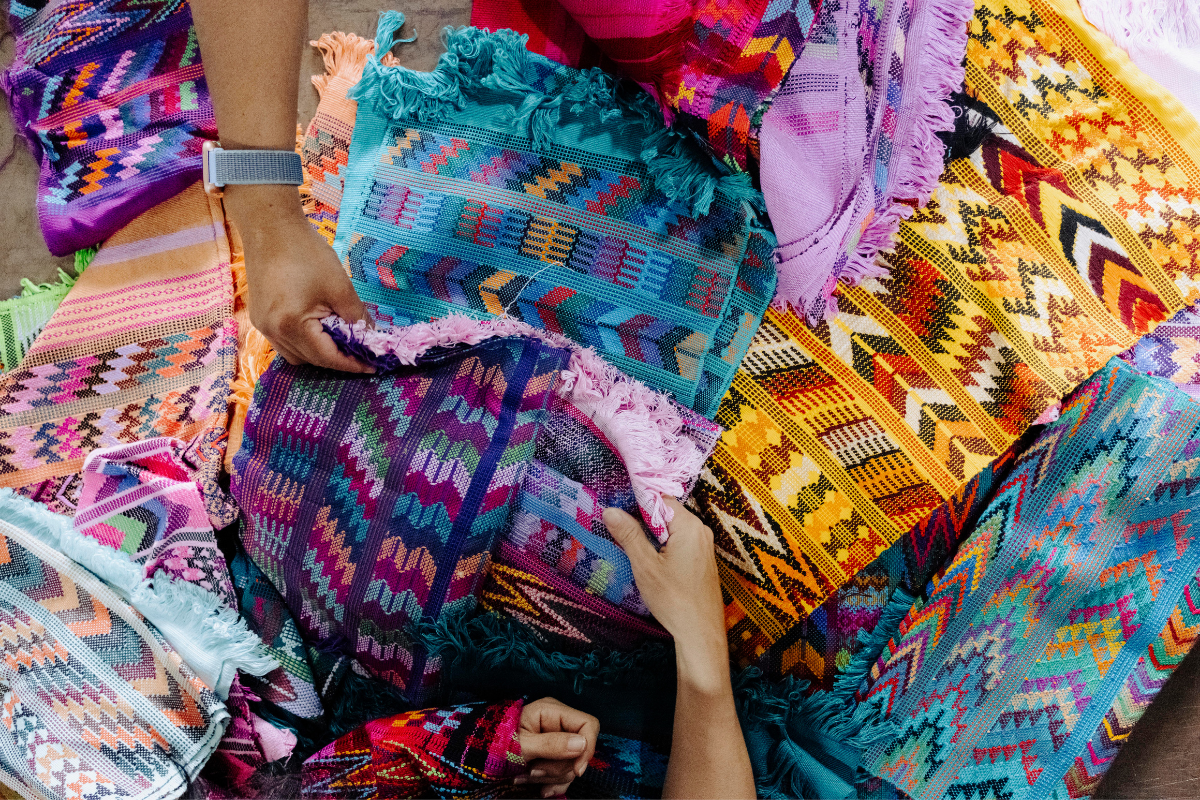 A close-up of hands holding and displaying vibrant, hand-woven textiles with intricate geometric patterns in purple, pink, blue, orange, and yellow.