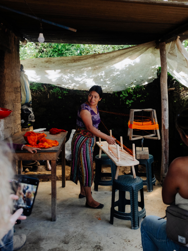 A woman stands under a rustic shelter, weaving a handcrafted wooden chair using a backstrap. She wears a purple patterned top and a colorful striped skirt.
