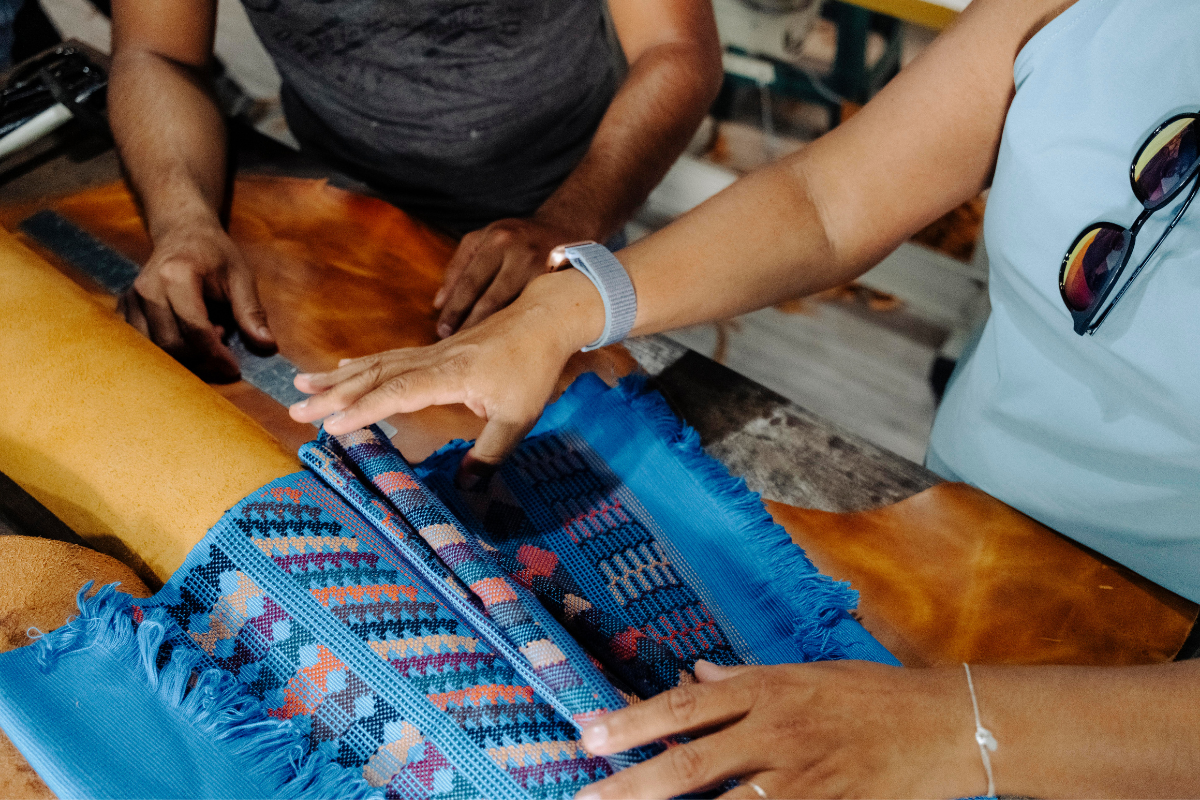 A close-up lifestyle shot of a person handling a vibrant blue and multicolor patterned woven textile, possibly a rug or tapestry, alongside another person at a wooden table.