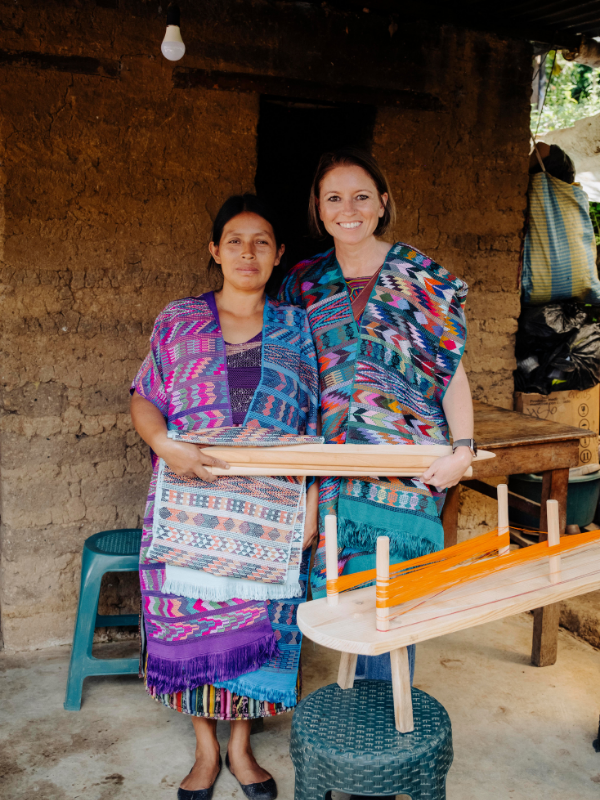 Two women standing in front of a rustic mud wall, wearing colorful patterned woven shawls and holding wooden furniture pieces in what appears to be a workshop or home setting.