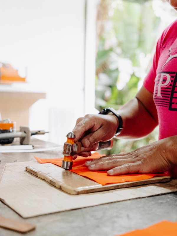A close-up lifestyle shot of a person's hands using a metal stamp with an orange handle to stamp an orange sheet of material onto a wooden board, set in a bright, softly blurred workshop environment.