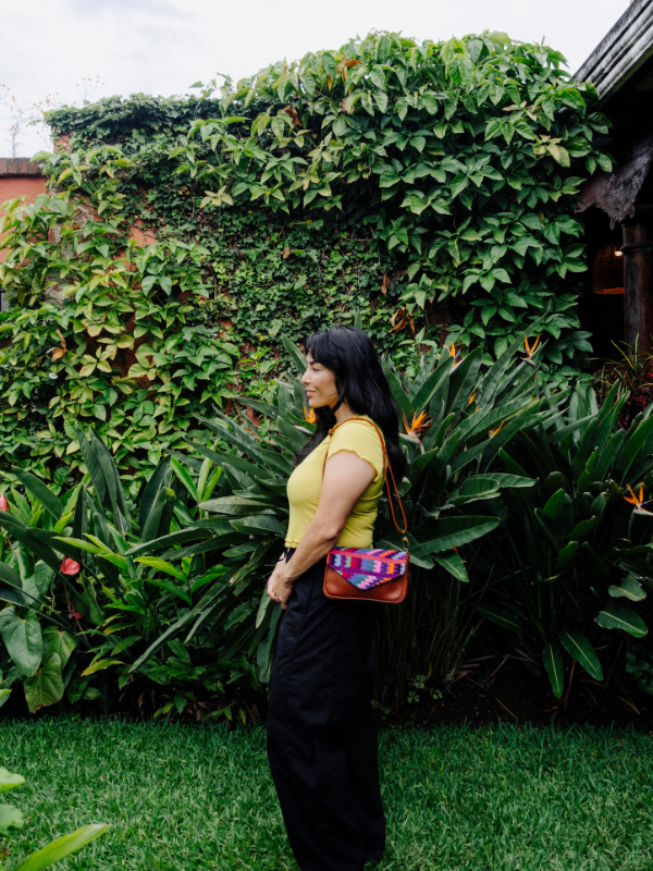 A woman stands in profile outdoors in front of a lush green ivy-covered wall, wearing a yellow top, black pants, and carrying a small brown crossbody bag with a colorful patterned fabric panel.