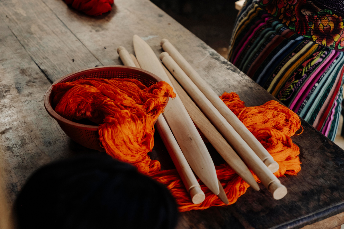 A close-up of a traditional weaving setup on a rustic wooden table, featuring bright orange yarn wound around wooden spindle tops and a colorful striped textile partially visible in the background.