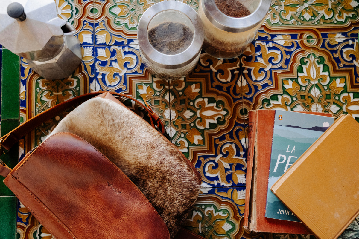 A brown leather shoulder bag with a brown fur trim, laid open on a colorful patterned tile surface next to a moka pot, glass jars, and books.