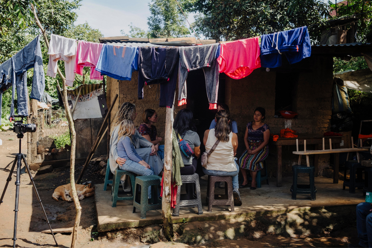 Several women sit on plastic stools under a makeshift shelter with a clothesline covered in colorful garments, with a camera on a tripod to the left in an outdoor rural setting.