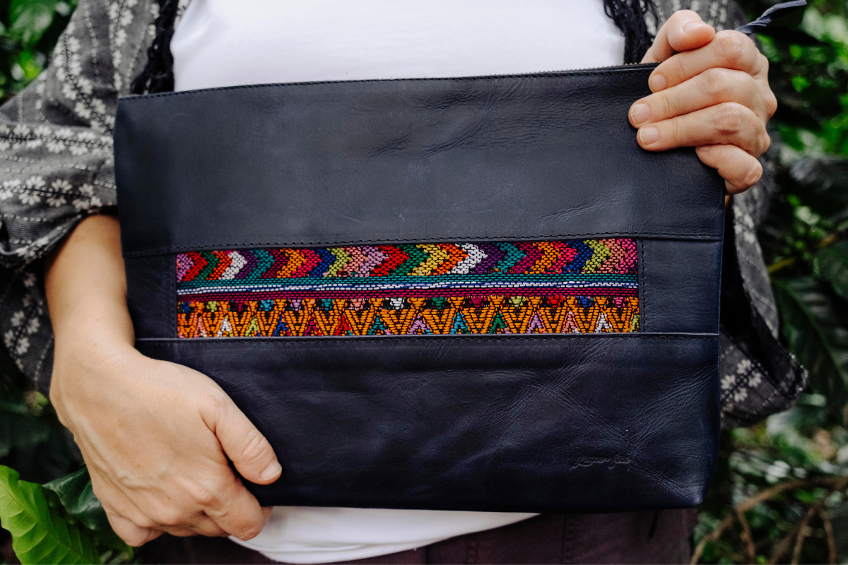 A person holds a black leather clutch bag with a colorful geometric embroidered band across the front, set against a blurred green outdoor background.