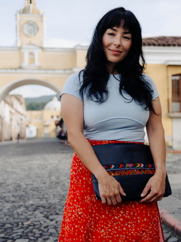A woman with long black hair stands outdoors in a cobblestone street setting, wearing a light blue short-sleeve top, a red floral maxi skirt, and holding a black clutch bag with colorful geometric embroidery.