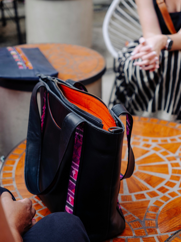 A black leather tote bag with a bright orange interior and purple patterned side accents, shown sitting on a mosaic outdoor table with people visible in the background and foreground.