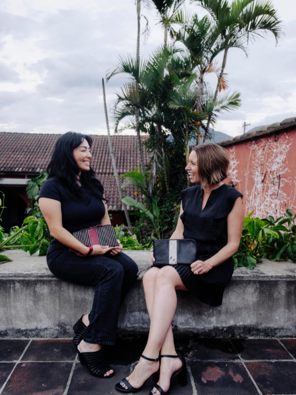 Two women sitting on a concrete ledge outdoors, both wearing black sleeveless dresses and black heels, holding black handbags in a tropical setting with palm trees and a terracotta wall.