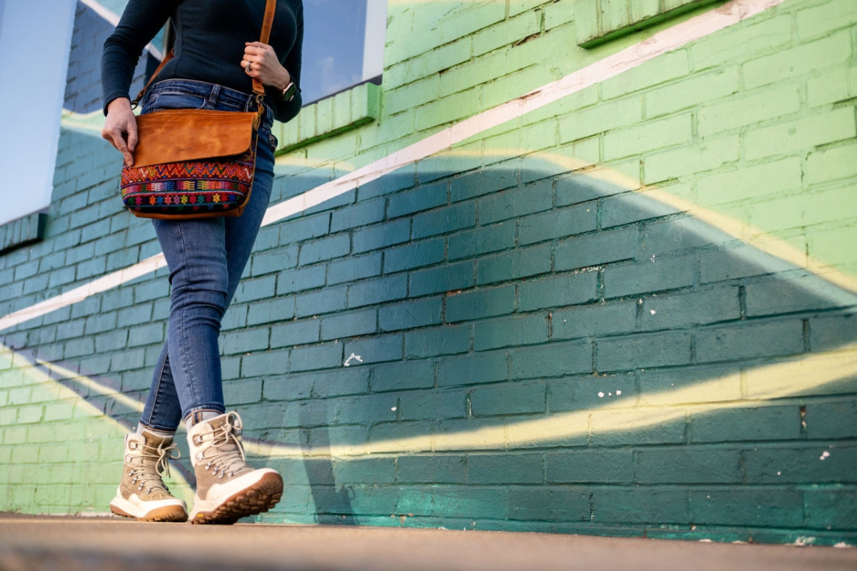 A person walking outdoors against a painted green brick wall, carrying a brown leather shoulder bag with colorful geometric embroidery and beige lace-up hiking boots.