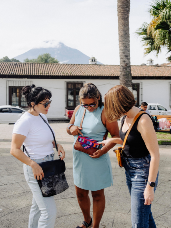 Three women standing outdoors on a paved street in front of a building and palm tree, looking at and holding a small patterned handbag. One woman wears a white t-shirt and light jeans, another wears a light blue dress, and the third wears a black tank top and jeans.
