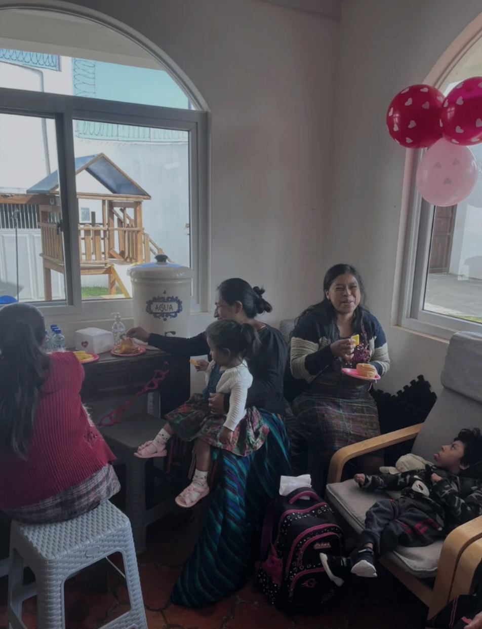 A family gathering inside a home with several women and children seated around a table, balloons on the wall, and a large window in the background.