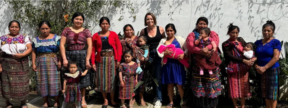 A group of women and children wearing traditional Mexican clothing, including colorful embroidered blouses, skirts, and dresses, posing together outdoors in front of a white wall.