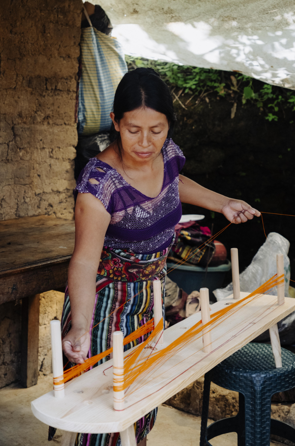 A woman in a purple and multicolored traditional woven dress sits at a wooden loom, actively weaving with orange cotton thread in a rustic outdoor workshop setting.