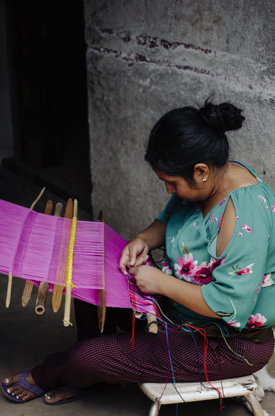 A woman sits on a small stool weaving a bright pink textile with wooden loom tools, wearing a teal floral top and dark patterned pants against a rustic concrete wall.