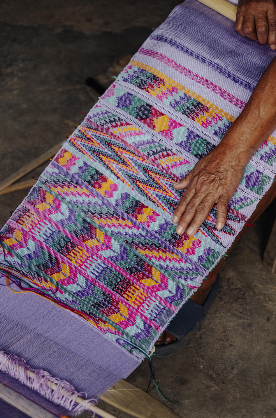 A close-up of a hand weaving a colorful geometric patterned textile on a loom, with purple and lavender tones dominating alongside pink, orange, green, and yellow accents.