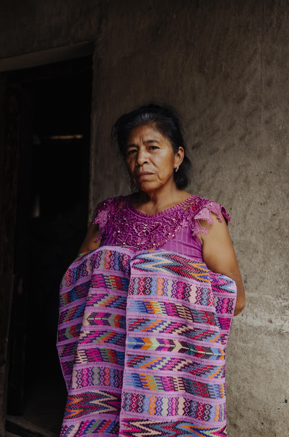 A woman wearing a vibrant pink and purple embroidered blouse and a matching patterned pink wrap draped over her shoulders, standing against a textured gray wall.