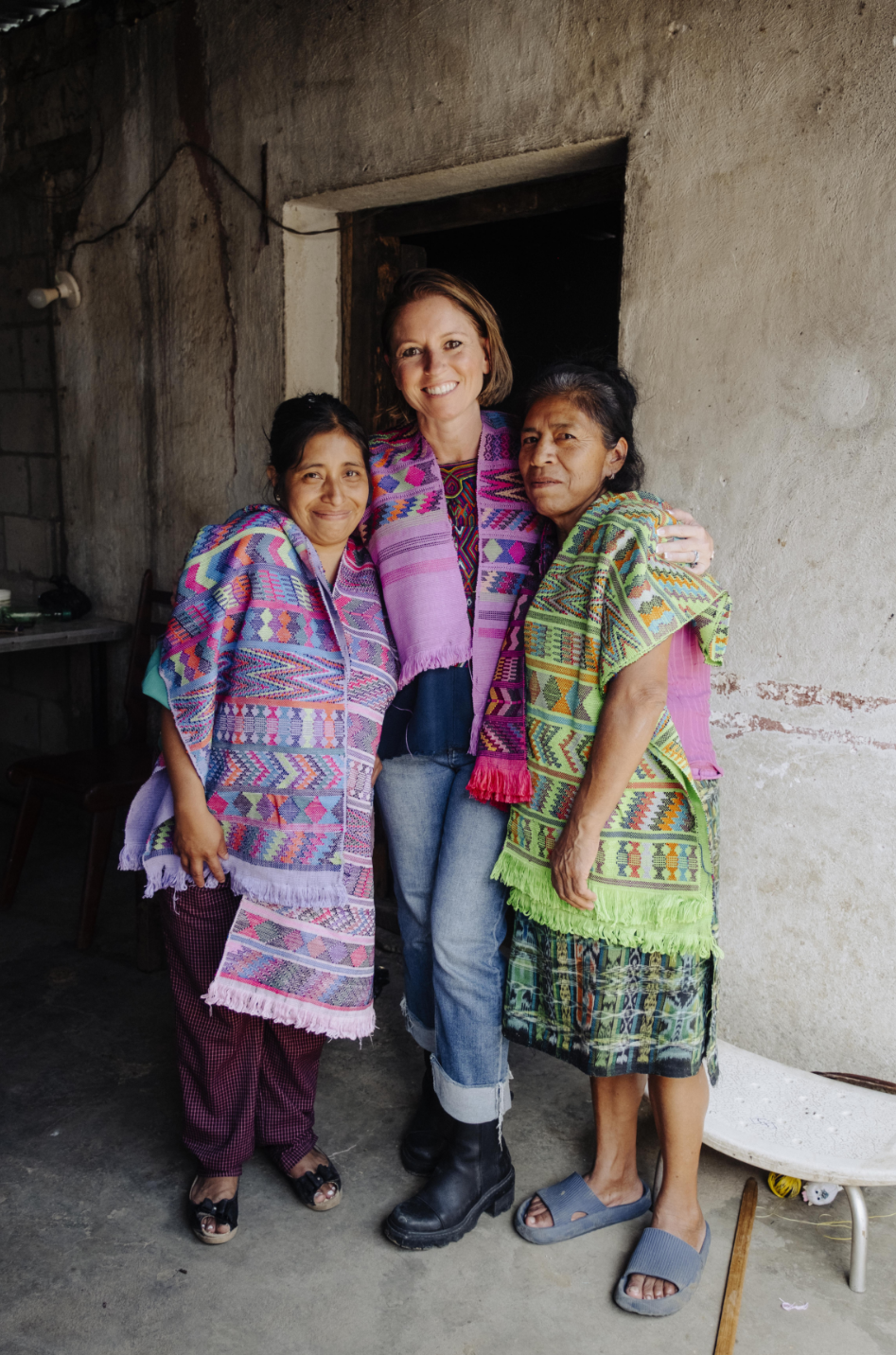 Three women standing together in front of a rustic concrete wall doorway, wearing colorful woven textiles and shawls. The central woman wears a pink and purple patterned shawl over a dark top and jeans, while the two women beside her wear brightly patterned woven garments in purple, green, and pink tones.