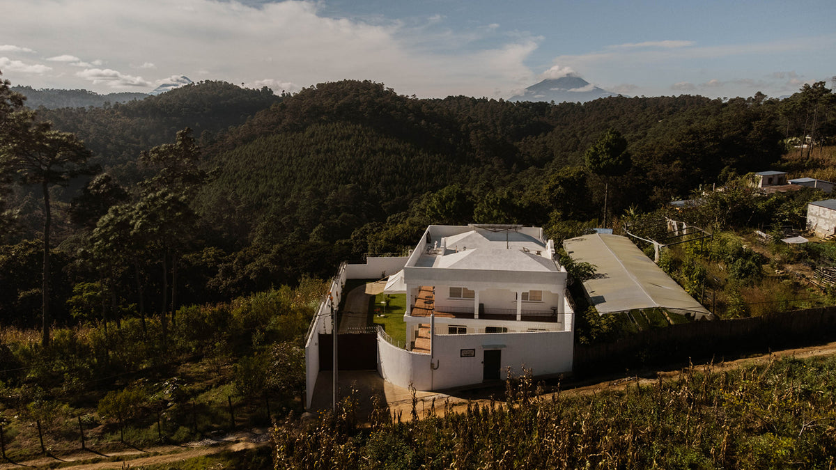 A modern white house with a curved roof sits on a hillside surrounded by dense green forest and mountains under a partly cloudy sky.